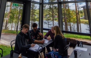 A group of researchers solve a science problem together in a glass building surrounded by trees, next to a river